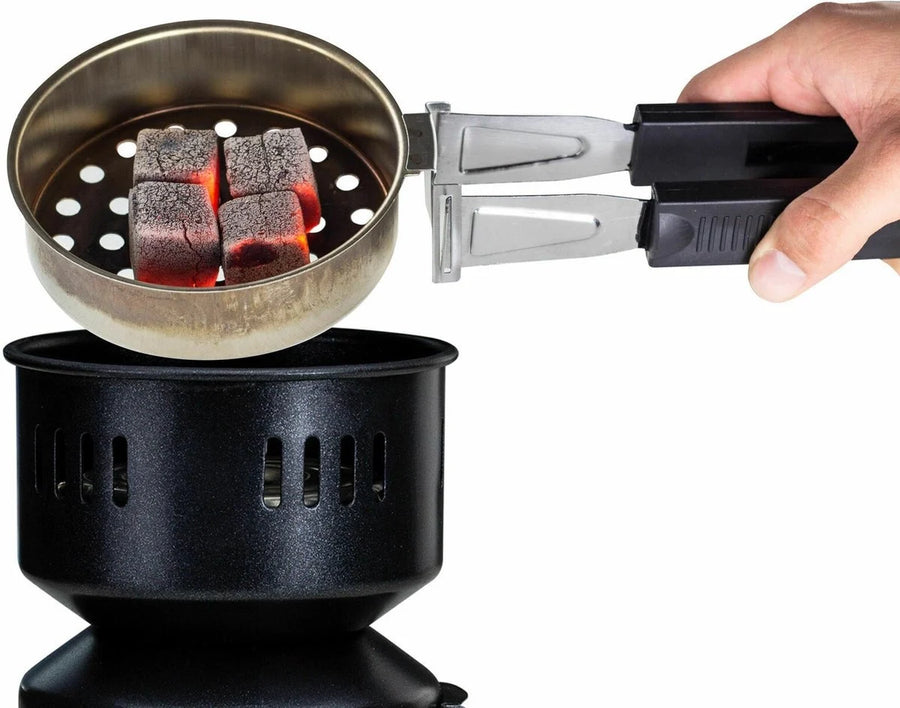 Een hand tilt met een tang een korf met vier gloeiende kokoskolen van de brander, de kubussen zijn rondom oranje aangegloeid en klaar voor de kop.
